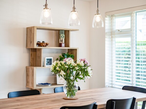 Uma área de refeições com mesa de madeira, cadeiras, um vaso de flores, luminárias pendentes e uma estante de madeira com objetos decorativos.
