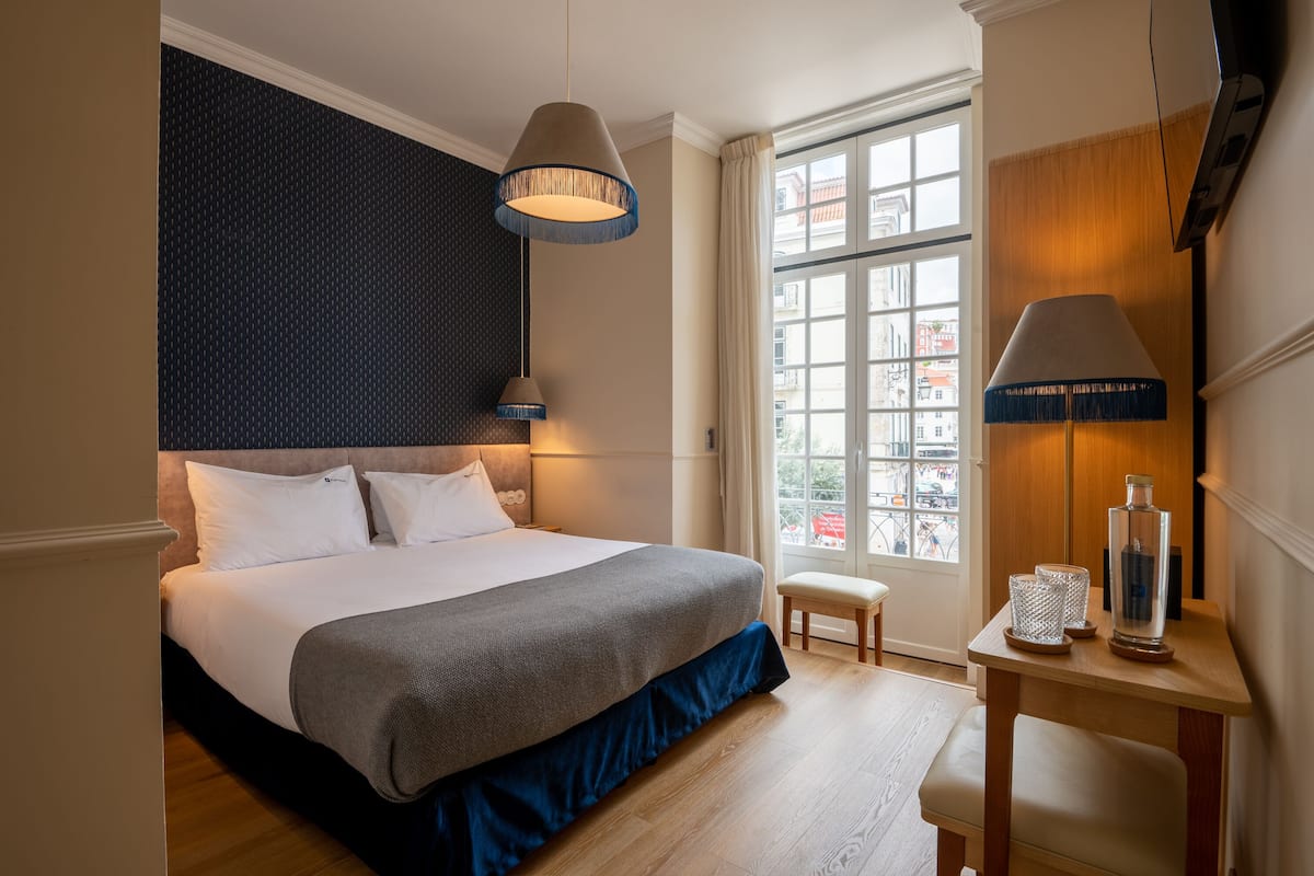 Une chambre d’hôtel avec un grand lit, une petite table avec une lampe et une fenêtre donnant sur des immeubles.