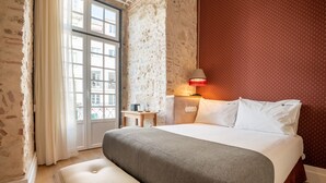 Hotelkamer met een groot bed, een bankje, een nachtkastje, een lamp en uitzicht op gebouwen door een raam.
