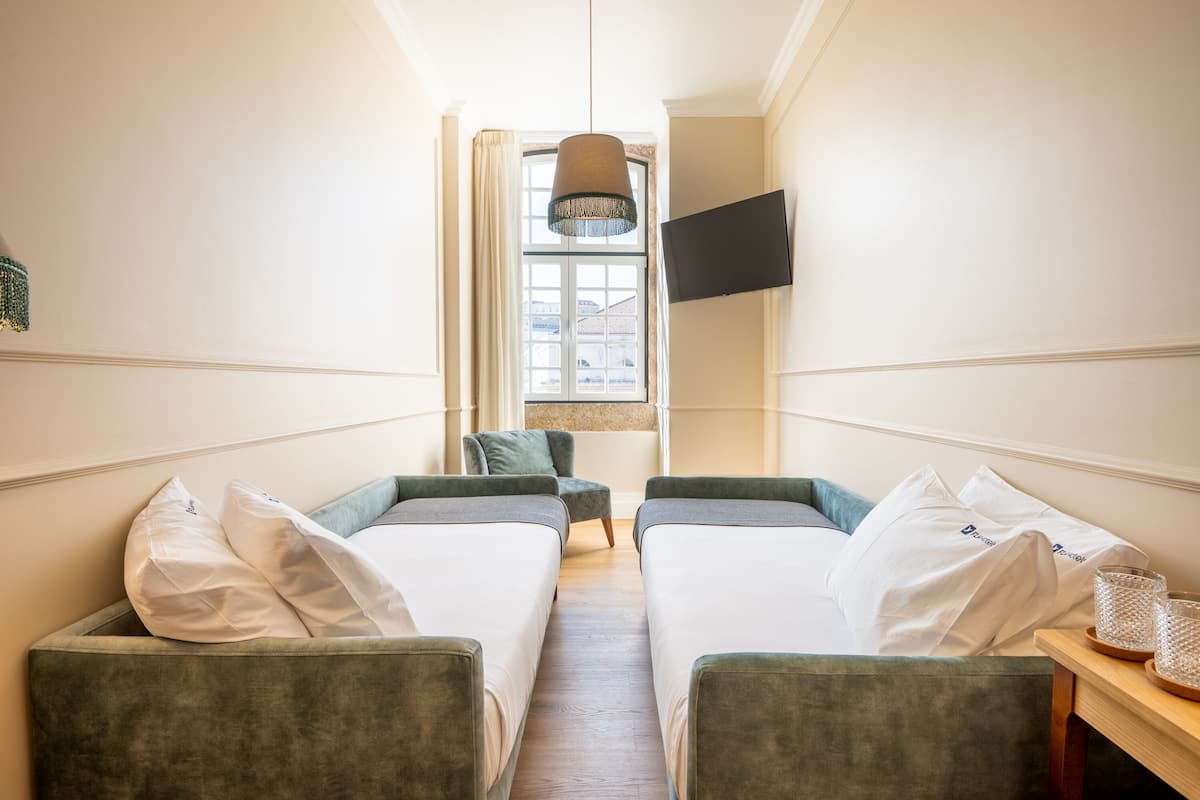 Une chambre d’hôtel avec deux lits simples, un téléviseur fixé au mur, une fenêtre avec des rideaux et une petite table avec des verres.