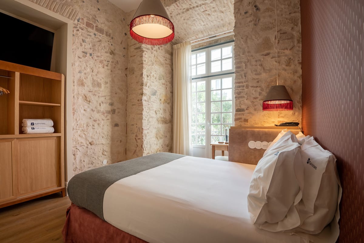 Une chambre d’hôtel avec un mur en pierre, une armoire en bois, un lit recouvert d’une couette blanche, une lampe rouge et une fenêtre donnant sur un paysage verdoyant.