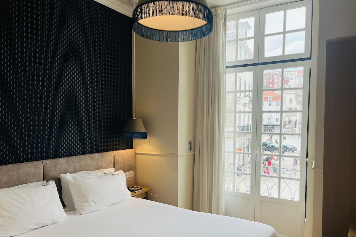Une chambre d’hôtel avec un grand lit, un mur à motifs sombres, une lampe suspendue et une fenêtre donnant sur des immeubles.
