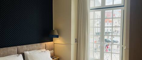 A hotel room with a large bed, a dark patterned wall, a hanging lamp, and a window with a view of buildings.