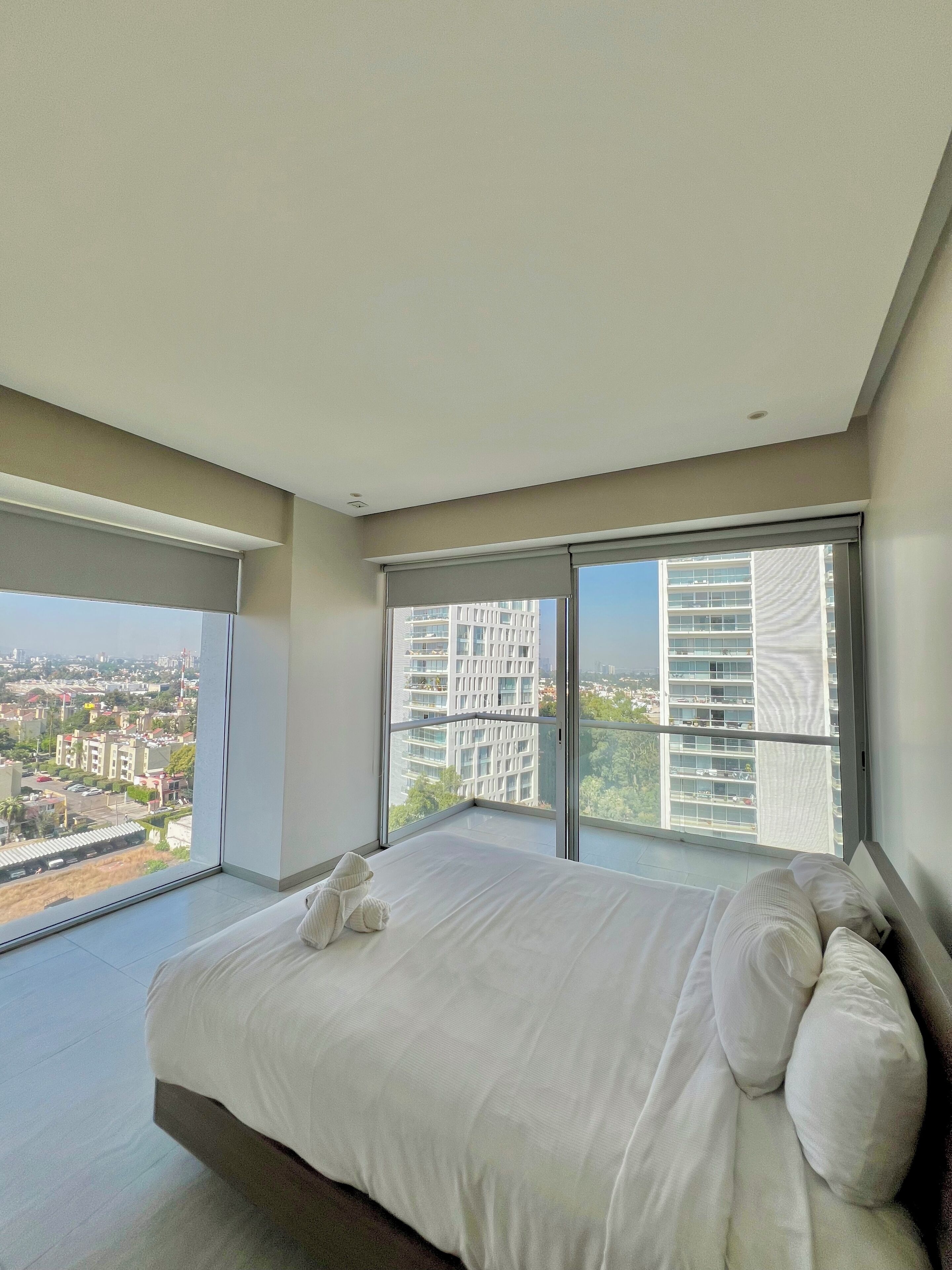 A modern bedroom with a large bed, a view of the city through floor-to-ceiling windows, and a balcony.
