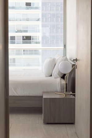 A bedroom with a bed, pillows, a bedside table with a lamp, and a window with a view of buildings.