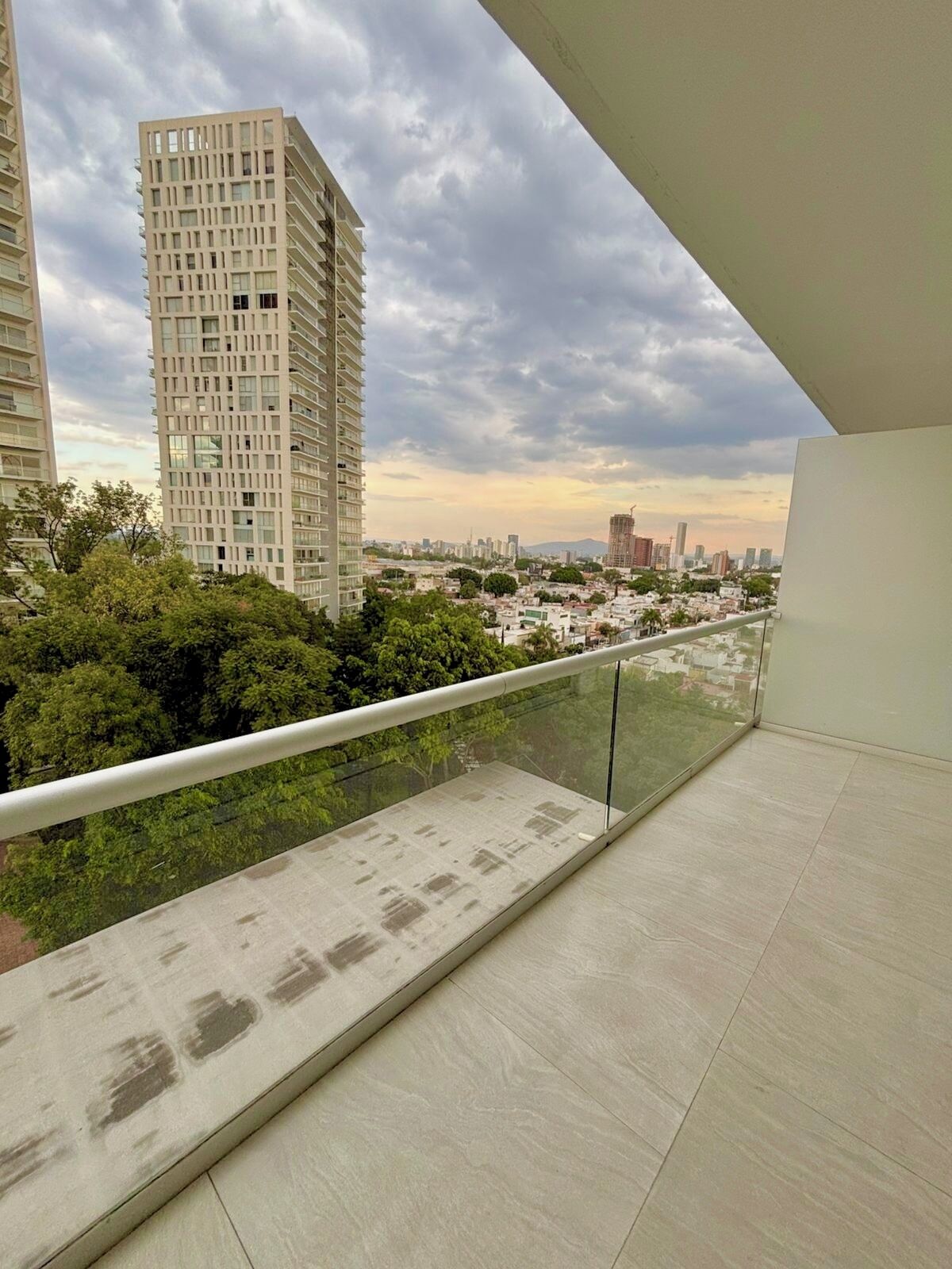 Een balkon met glazen balustrade, met uitzicht op een stadsgezicht met hoge gebouwen en groen.