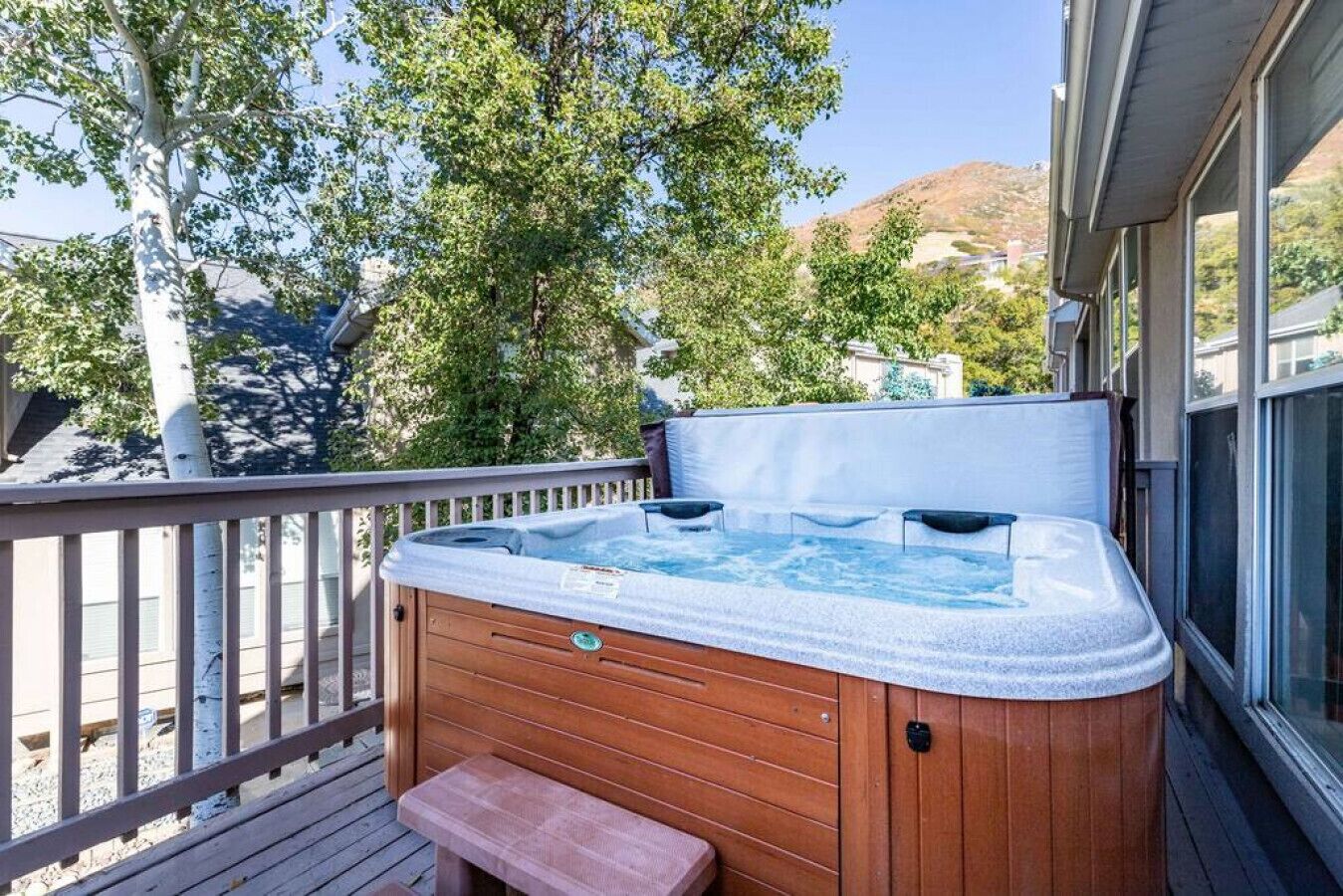 A hot tub on a wooden deck with a bench, surrounded by trees and a mountain view.