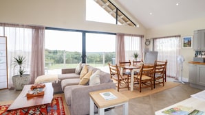 A spacious living area with a sofa, dining table, and chairs, large windows with curtains, and a view of greenery outside. - The Lodge (Stranraer)