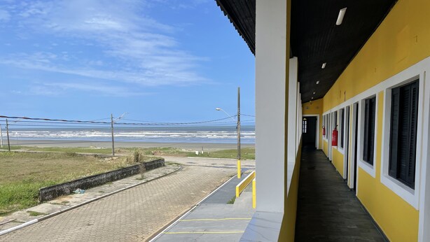 A coastal view from a building with a yellow exterior, a paved walkway, and a clear sky. - Suítes Temporada Favorita (Peruíbe)