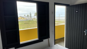 A room with a bed, two windows with shutters, a view of a grassy area and buildings, and a door with a number plate. - Suítes Temporada Favorita (Peruíbe)