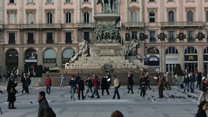 Una plaza pública con una estatua central, rodeada de edificios históricos de varios pisos y grandes ventanales.