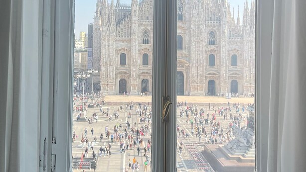 Vista a través de una ventana de un edificio imponente con arquitectura elaborada, una plaza concurrida al frente y un cielo despejado.