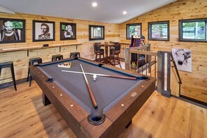A pool table in a room with wooden walls, a bar area with stools, and framed artwork on the walls.