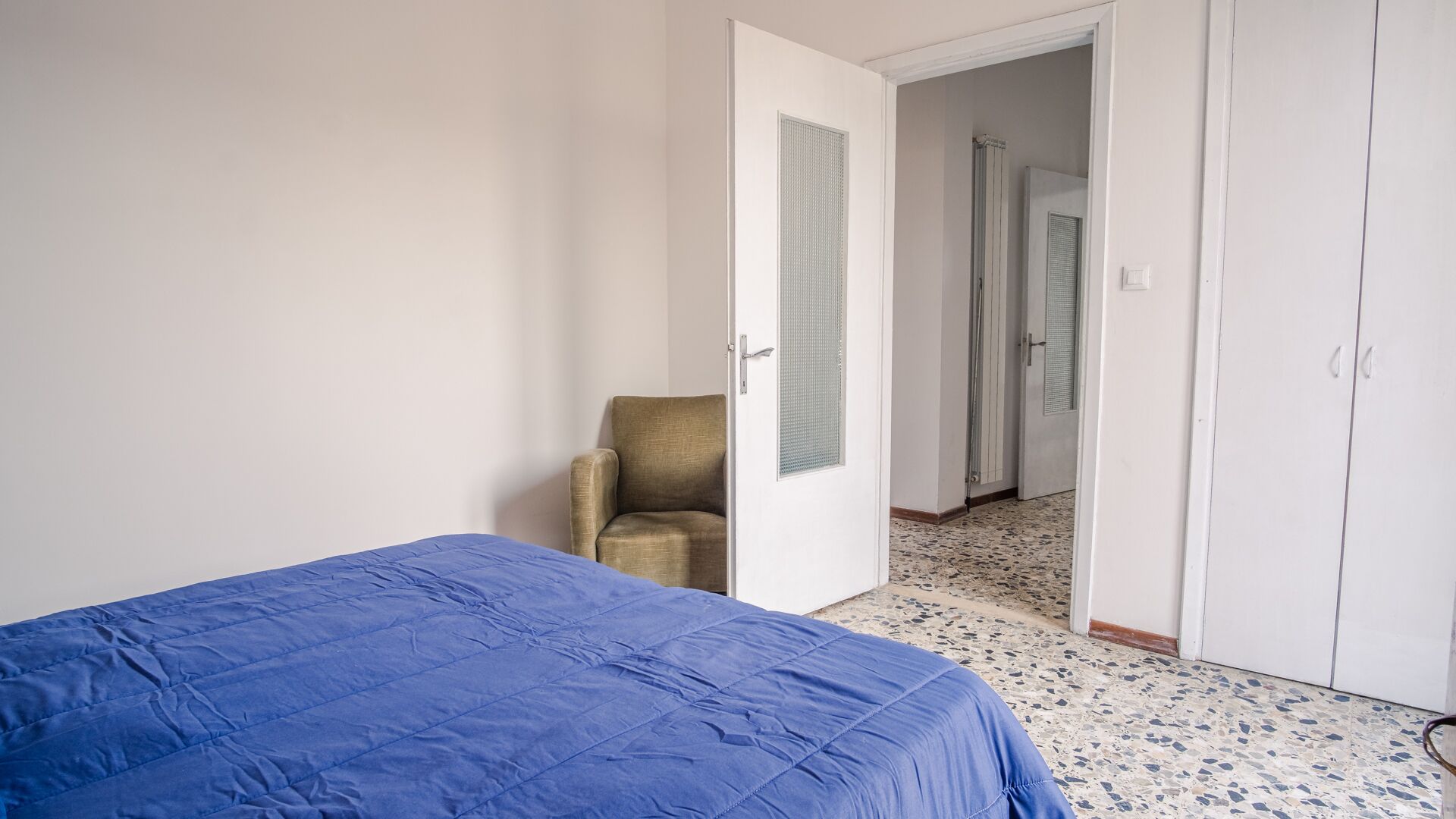 A bedroom with a bed, a chair, a wardrobe, and a door leading to another room.