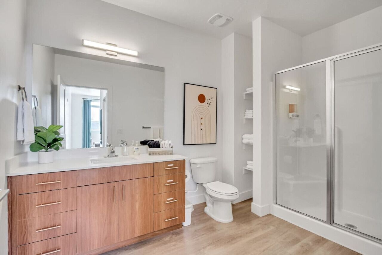 A modern bathroom with a wooden vanity, a toilet, a glass shower enclosure, and a framed artwork on the wall.