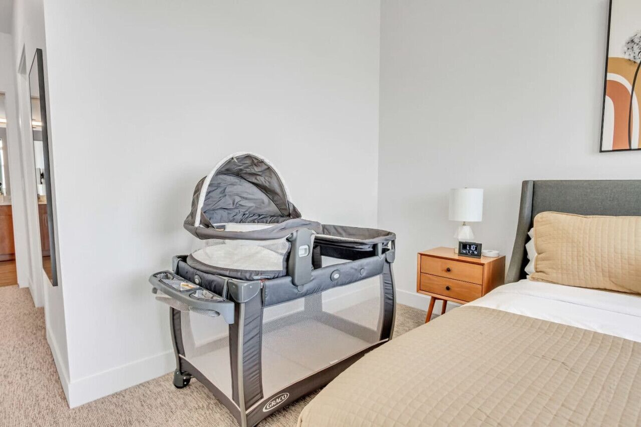 A room with a bed, a nightstand with a lamp, a baby crib, and a wall-mounted artwork.