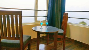 Un balcón con una mesa y dos sillas, una ventana con cortinas azules y un vaso con líquido azul sobre la mesa.