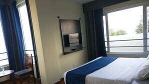 Habitación de hotel con cama, televisor montado en la pared, escritorio con silla y ventana con cortinas.
