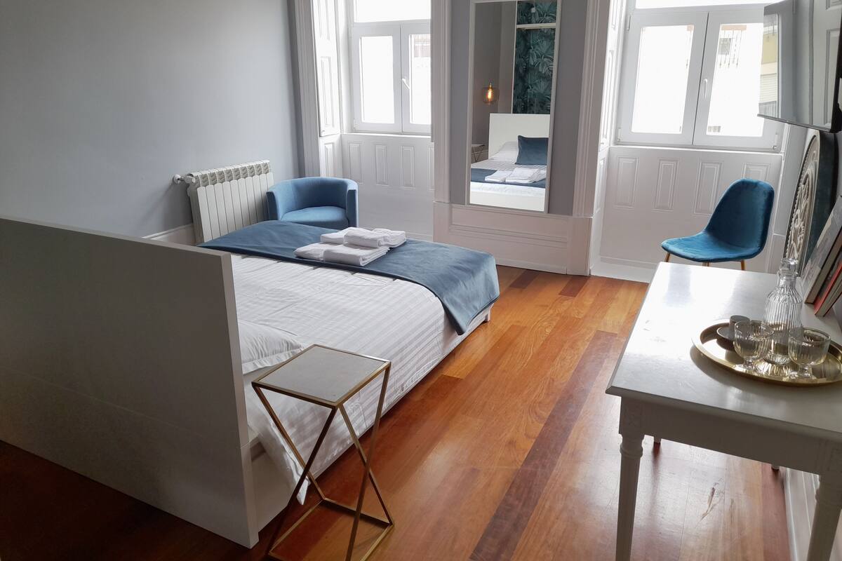 A bedroom with a bed, a blue chair, a small table with a tray, and a window with a view of another room.