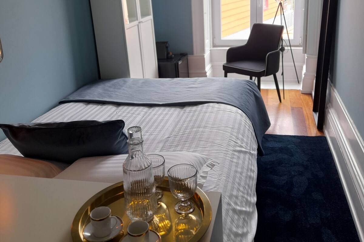 A bedroom with a bed, a bedside table with a tray of glasses and a bottle, a chair, a wardrobe, and a window with a view of the outside.