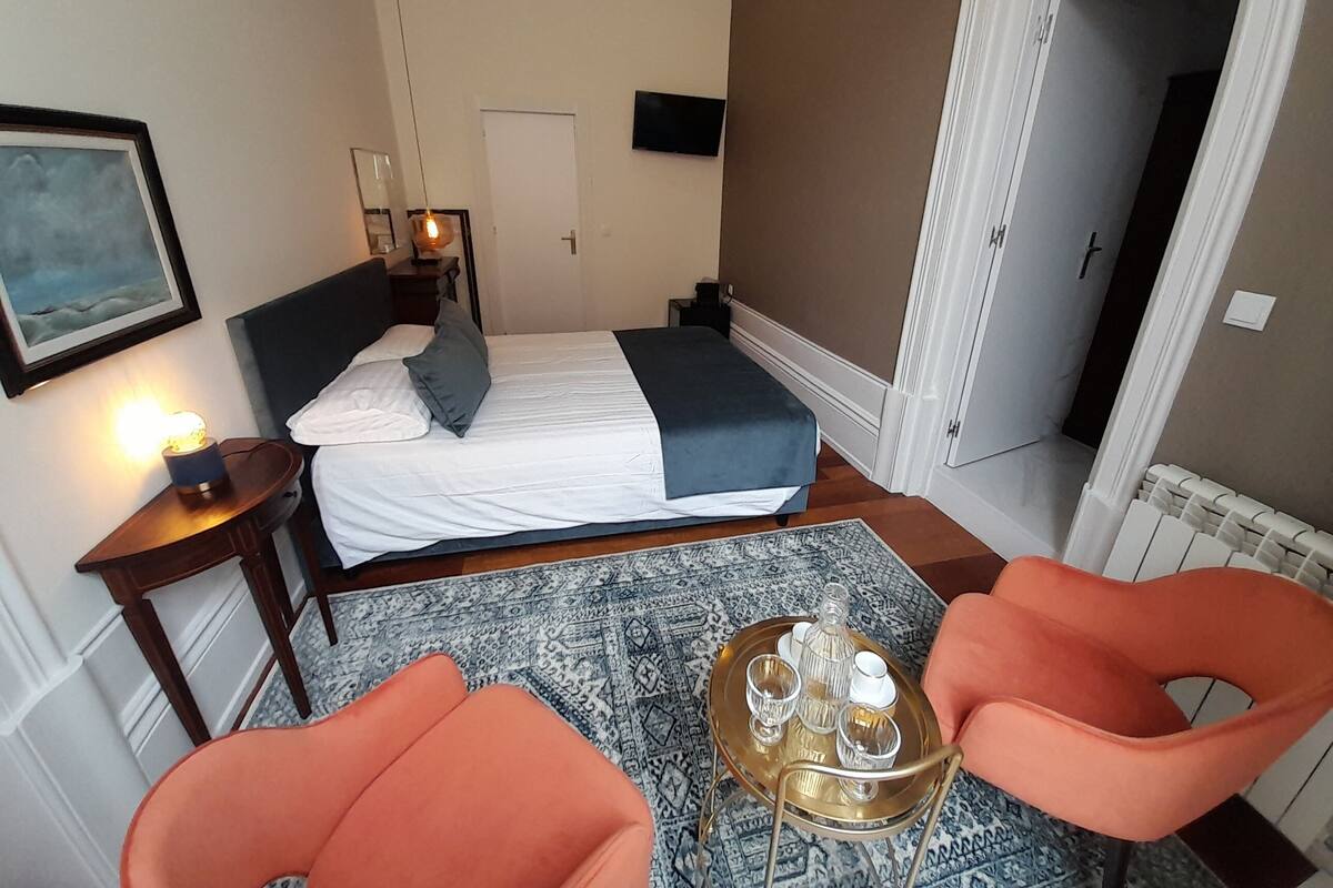 A hotel room with a bed, bedside table with a lamp, a small table with a tray of glasses and a bottle, and two orange chairs.