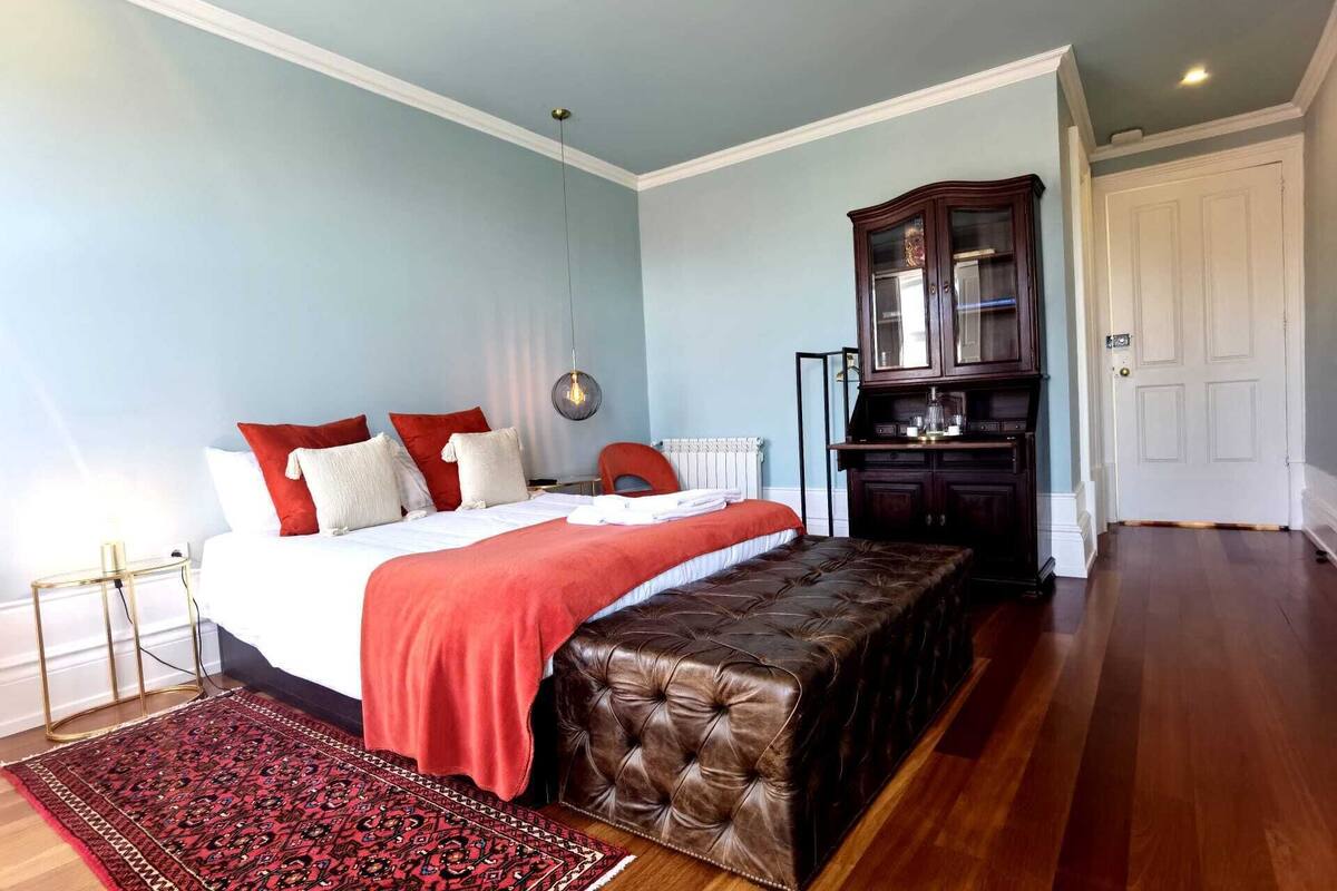 A bedroom with a bed, a red and white bedspread, a brown ottoman, a wooden cabinet, and a red rug.