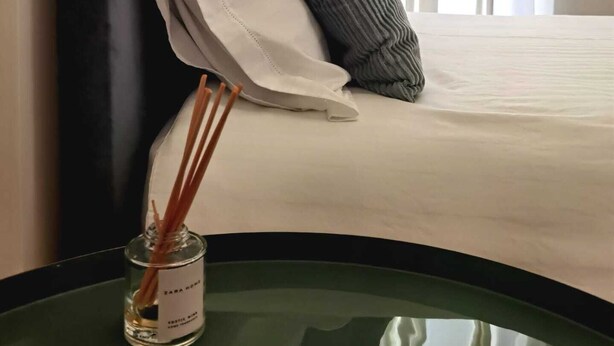 A neatly made bed with a white and gray pillow, a dark headboard, and a small table with a bottle of perfume. - Monsenhor52 (Porto)