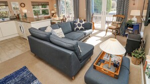 A living room with a grey sofa, a coffee table with books and a lamp, a dining area with a table and chairs, and a kitchen with a washing machine. - 1 Hornbeam Lodge (Narberth)