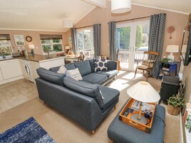 A living room with a grey sofa, a coffee table with books and a lamp, a dining area with a table and chairs, and a kitchen with a washing machine.