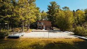 A two-story house with a deck and a sandy driveway, surrounded by trees.
