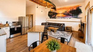 A modern kitchen with stainless steel appliances, a wooden dining table with a plant, a living area with a sofa, and a mural of a sunset. - New! Spider Lake| Hot Tub| Sauna| Beach| No Bunks| (Traverse City)