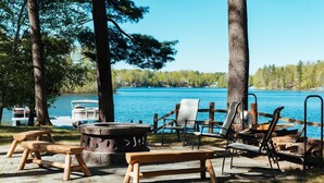 A lakeside seating area with a fire pit, benches, and chairs, surrounded by trees and a view of the lake. - Fishing-Spider Lake-Sauna-Balcony-Family Friendly (Traverse City)