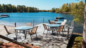 A wooden deck with tables and chairs overlooking a lake, surrounded by trees. - Spider Lake  | Bonfires | Beach | Hot Tub l Sauna (Traverse City)