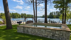A stone hot tub surrounded by a stone wall, a wooden deck, and a view of a lake with trees. - Spider Lake  | Bonfires | Beach | Hot Tub l Sauna (Traverse City)