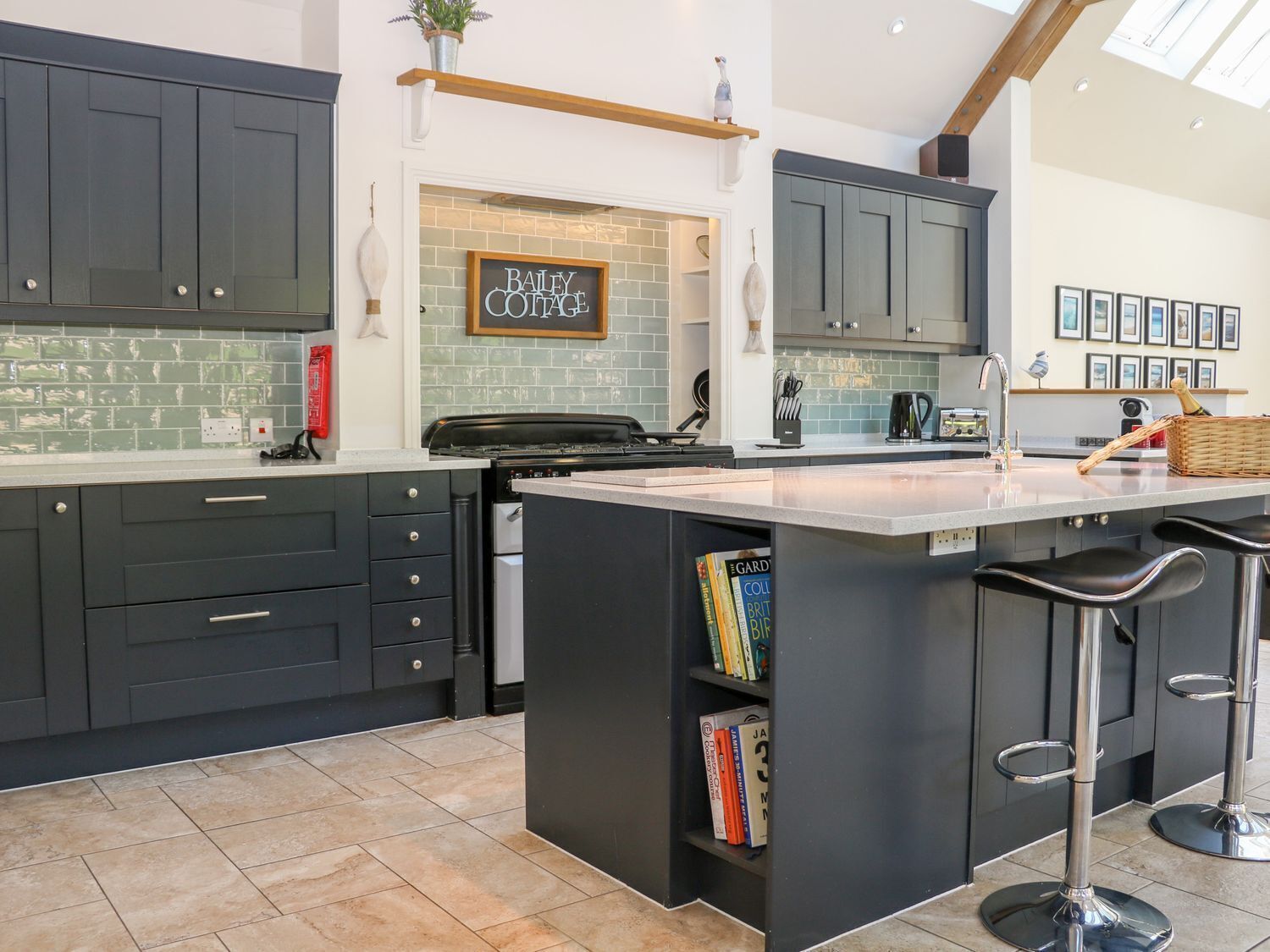 A modern kitchen with dark cabinets, a central island with bar stools, a backsplash with a 'Bailey Cottage' sign, and a tiled floor.
