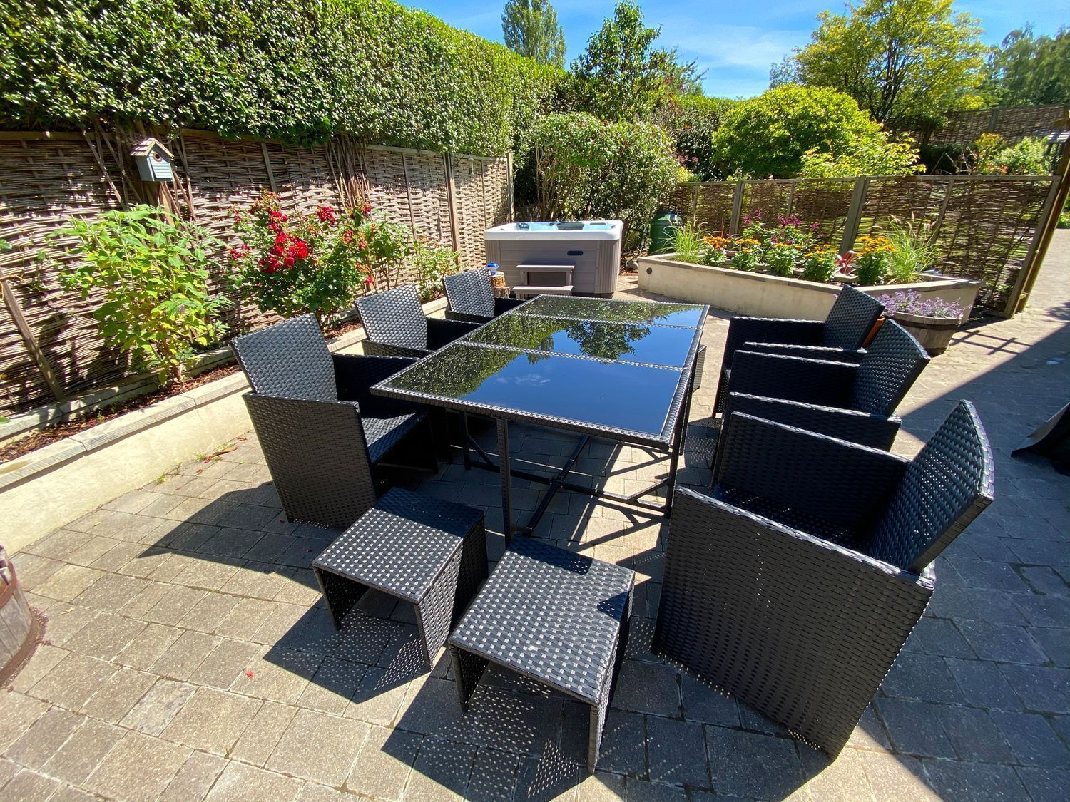 Eine Terrasse im Freien mit einem Tisch aus Glasplatten und Korbstühlen, umgeben von einem Garten mit verschiedenen Pflanzen und einem Holzzaun.