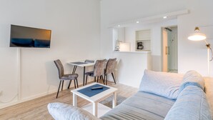 A modern living room with a sofa, a coffee table, and a TV mounted on the wall. There is a dining area with chairs and a small table. - Alte Teestube 55 , Weststrand Wifi Kostenlos Sofortige Buchung Balkon Modernes Ein - Raum - Appartement für bis zu 2 Personen (Norderney)