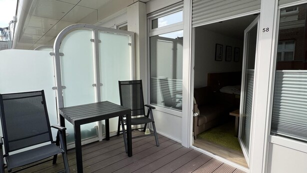 A covered outdoor area with a table and chairs, a sliding glass door, and a view of the interior. - Alte Teestube 55 , Weststrand Wifi Kostenlos Sofortige Buchung Balkon Modernes Ein - Raum - Appartement für bis zu 2 Personen (Norderney)