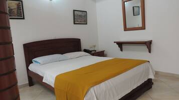 A bedroom with a wooden bed, white bedding, a mustard-colored throw blanket, a nightstand with a phone, and a small wooden shelf.