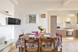 A dining area with a table set for two, a TV mounted on the wall, and a kitchen in the background.