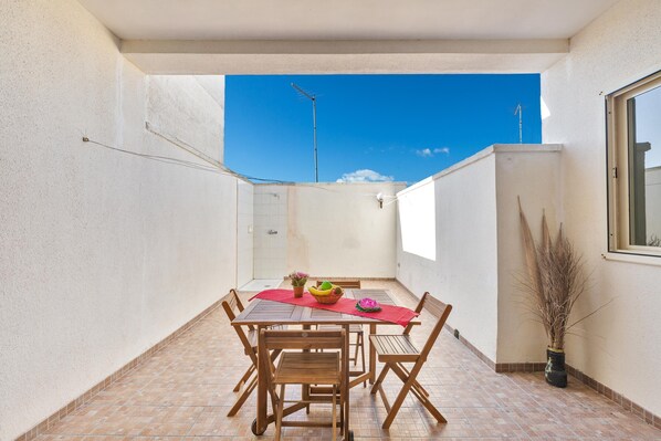 A small, enclosed patio area with a tiled floor, a wooden table with chairs, a red tablecloth, and a potted plant.
