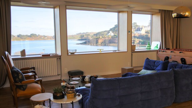 A living room with a large window overlooking a body of water, a blue sofa, a wooden chair, a small round table with a plant, and a radiator. - Villa Avec vue Mer, Piscine Chauffée (Landéda)
