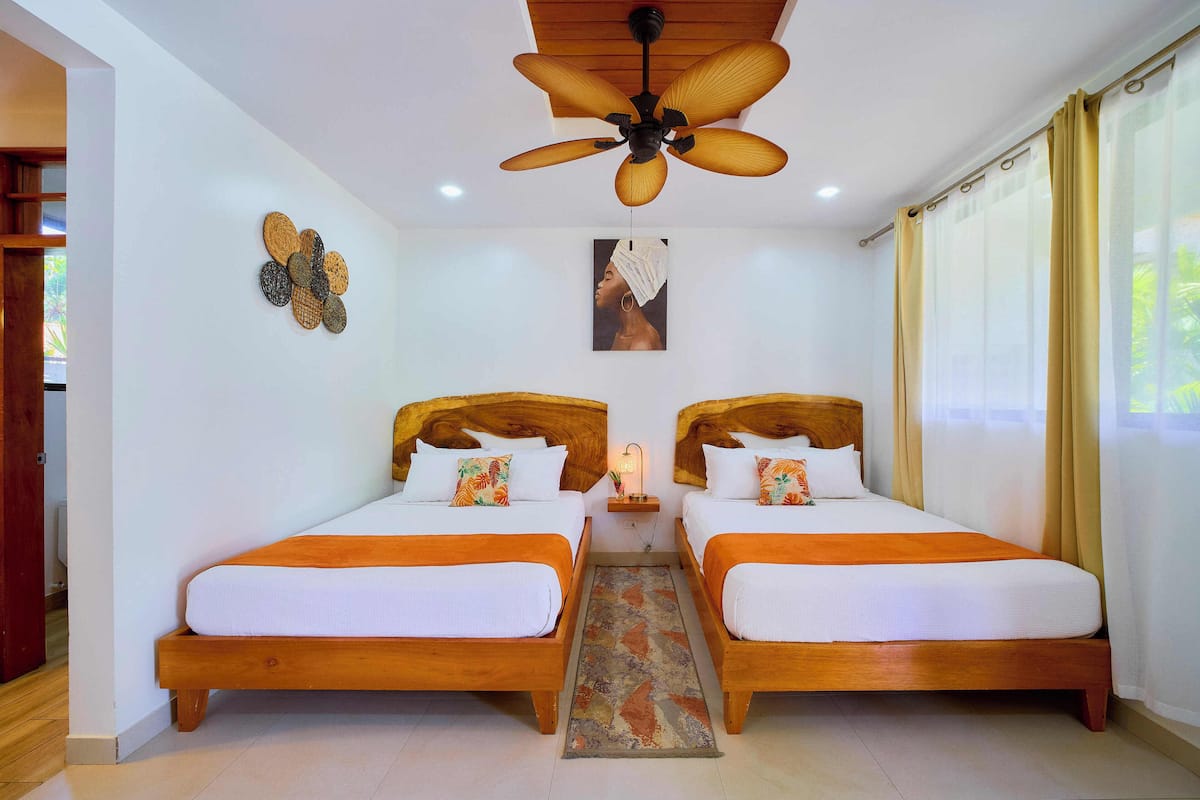 Une chambre d’hôtel avec deux lits en bois, un ventilateur de plafond, une petite table avec une lampe et un élément décoratif mural.