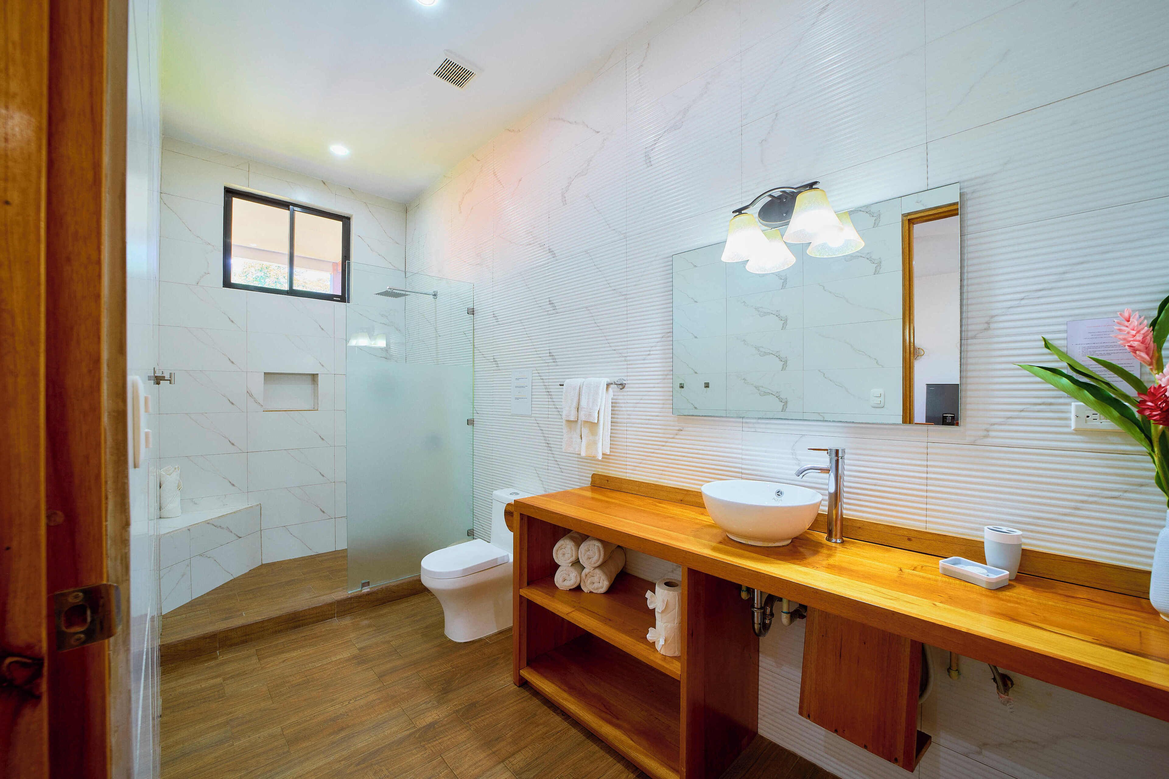 A modern bathroom with a wooden vanity, white marble tiles, a large mirror, and a shower area.