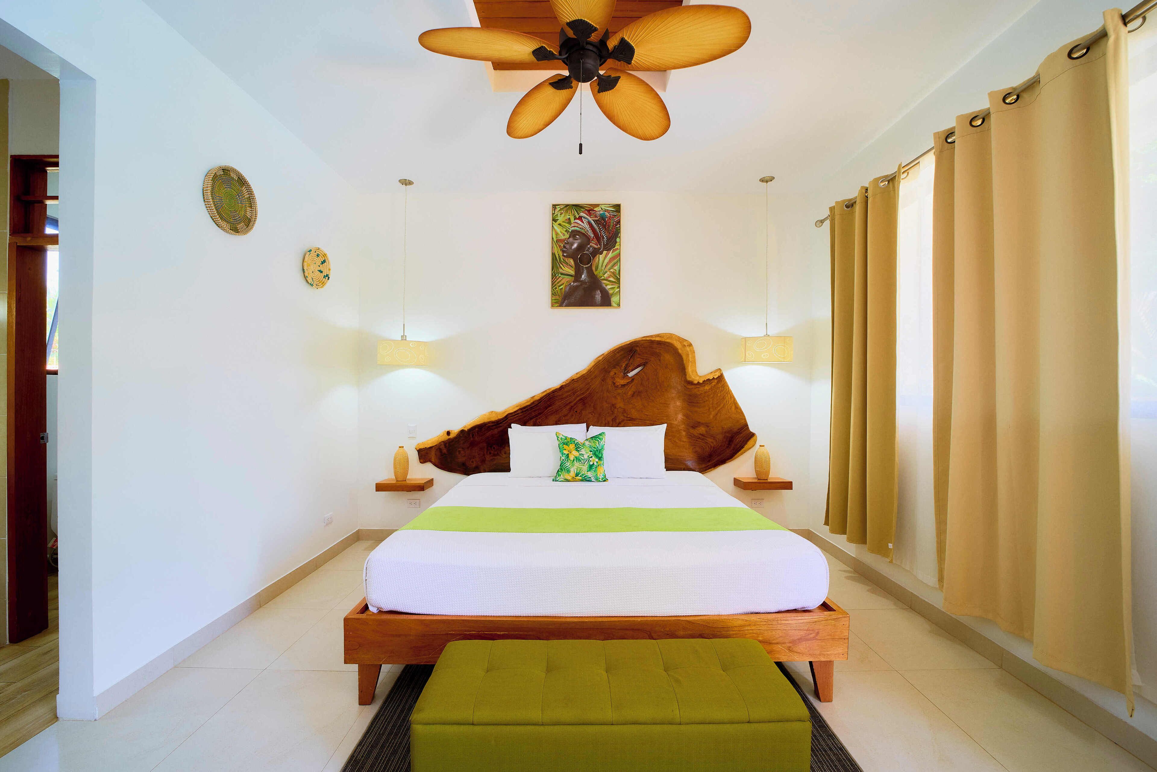 Quarto com cabeceira de madeira, cama com roupa de cama branca, um banco verde, ventoinha no teto e uma pintura na parede.