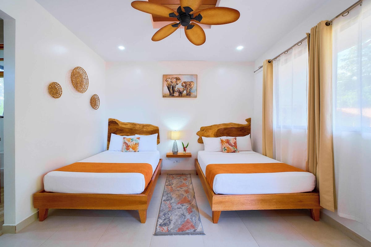 Une chambre d’hôtel avec deux lits en bois, un ventilateur de plafond, un tableau au mur et une fenêtre avec des rideaux.