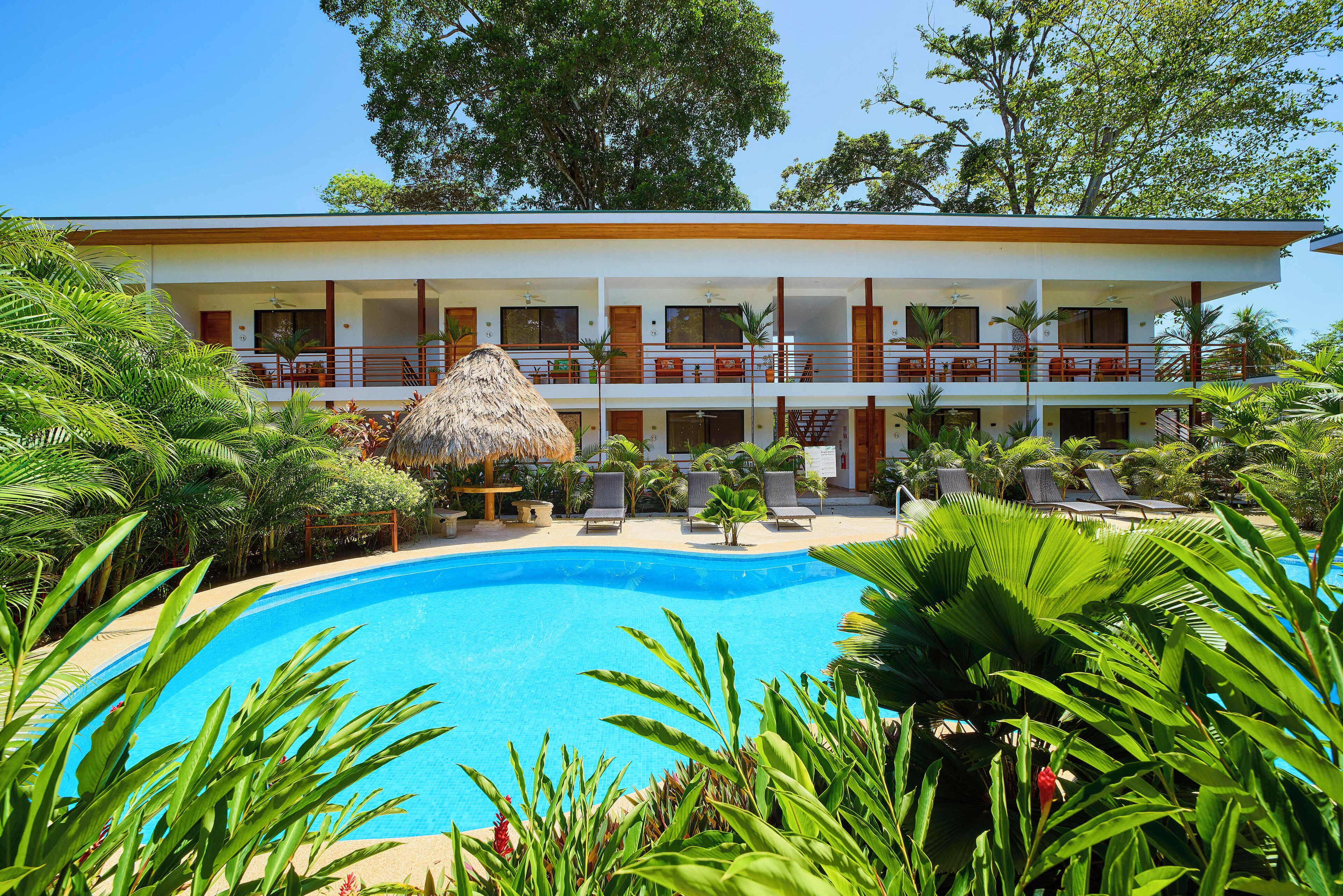 A hotel with a pool surrounded by lush greenery and a thatched-roof structure.