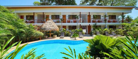 A hotel with a pool surrounded by lush greenery and a thatched-roof structure.