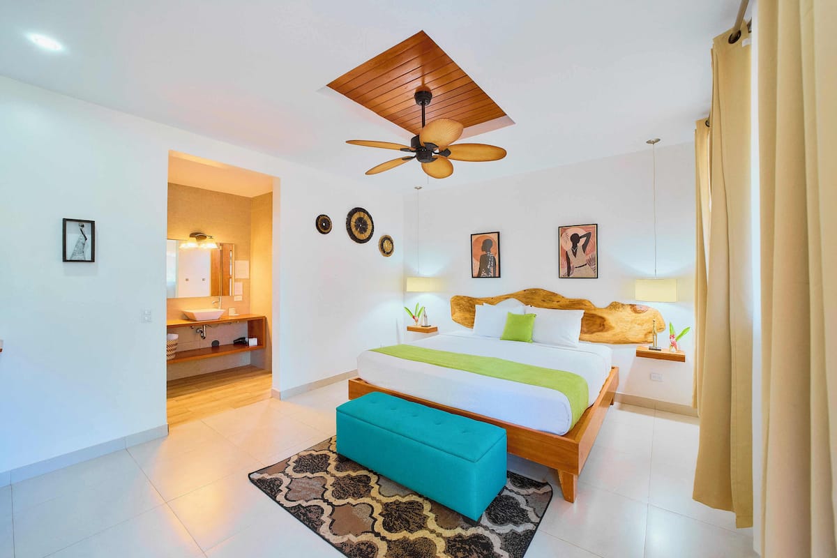 Une chambre d’hôtel moderne avec un ventilateur de plafond en bois, un grand lit recouvert d’un plaid vert, une tête de lit en bois et une petite table de chevet avec une lampe.