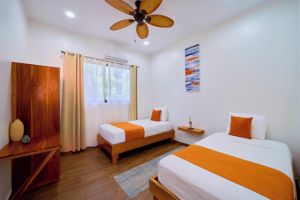 Une chambre d’hôtel avec deux lits, des meubles en bois, un ventilateur de plafond et une fenêtre avec des rideaux.